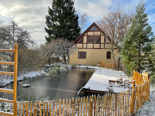 Ein Gartenteich vor einem Haus mit Fachwerk. Auf dem Steg liegt Schnee.