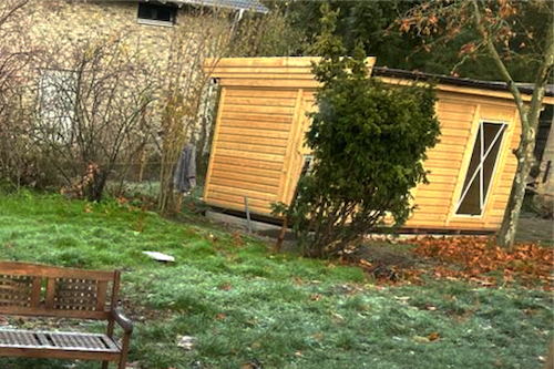 Hinter einem Baum steht eine Sauna aus Holz.