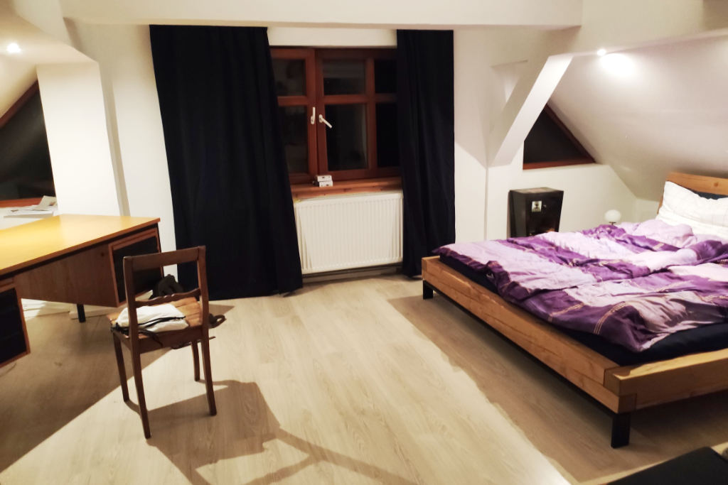 Ein Zimmer, links steht ein Schreibtisch mit Stuhl, rechts ein Bett mit lilafarbenem Bettbezug