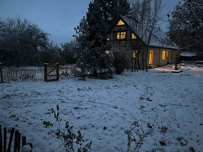 Vor dunklem Himmel liegt eine schneebedeckte Fläche. In Hintergrund ist ein Haus mit erleuchteten Fenstern zu sehen.