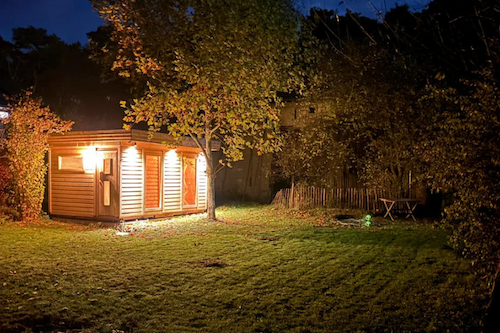 Eine beleuchtete Holzhütte steht auf einer Wiese. Es ist dunkel.