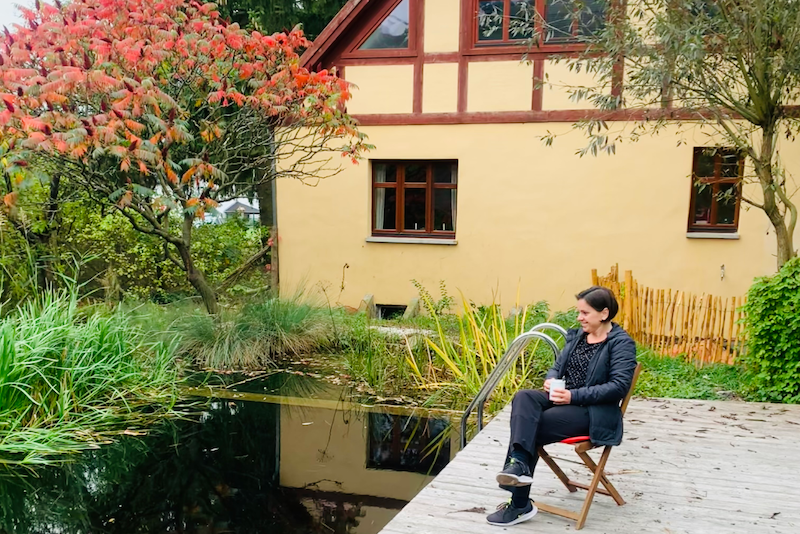 Vor einem kleinen See steht ein Haus. Rechts sitzt auf einem Steg eine Frau