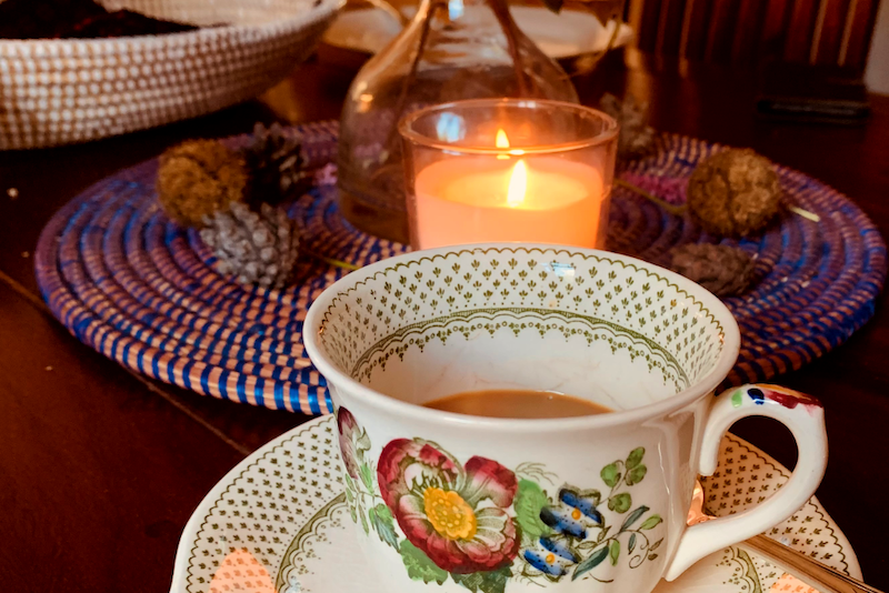 Auf einem dunkelbraunen Tisch steht eine Teetasse. Im Hintergrund brennt eine Kerze.